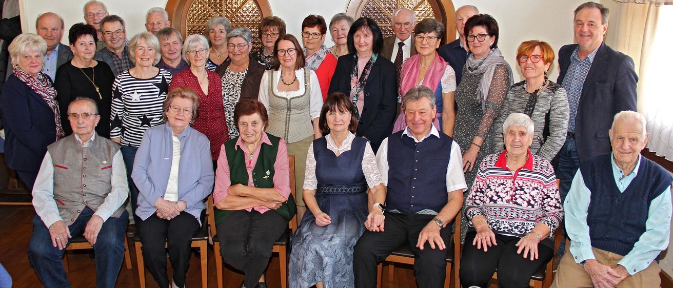 Eine Gruppe von Menschen, überwiegend ältere Personen, posiert für ein Foto in einem Raum mit Holzwänden und Bogenfenstern. Sie sitzen und stehen, tragen Brillen, Halsketten und teilweise formelle Kleidung.