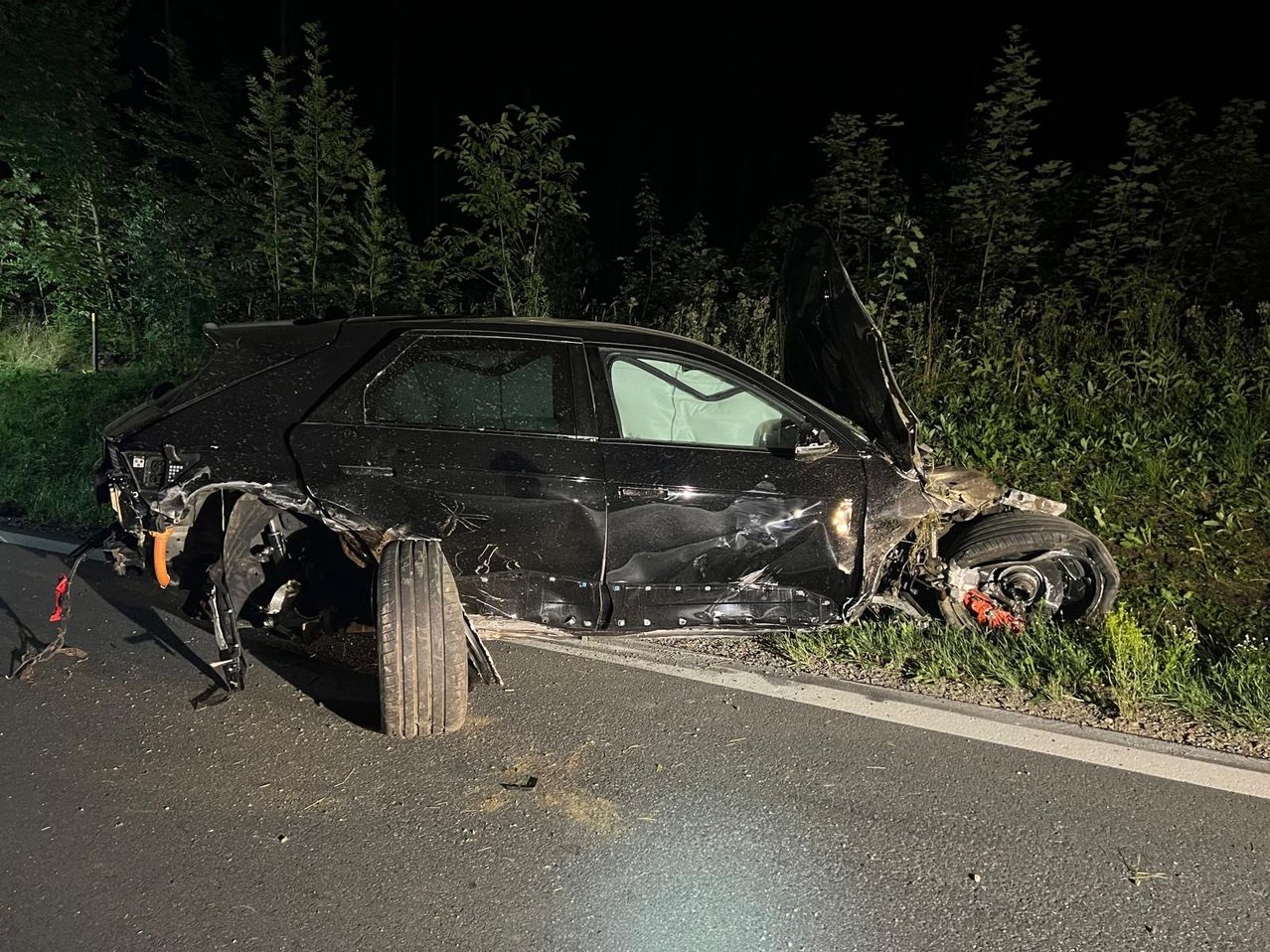 Ein schwarzer Wagen ist nachts an der Straßenseite schwer beschädigt, mit starker Front- und geöffneter Motorhaube.