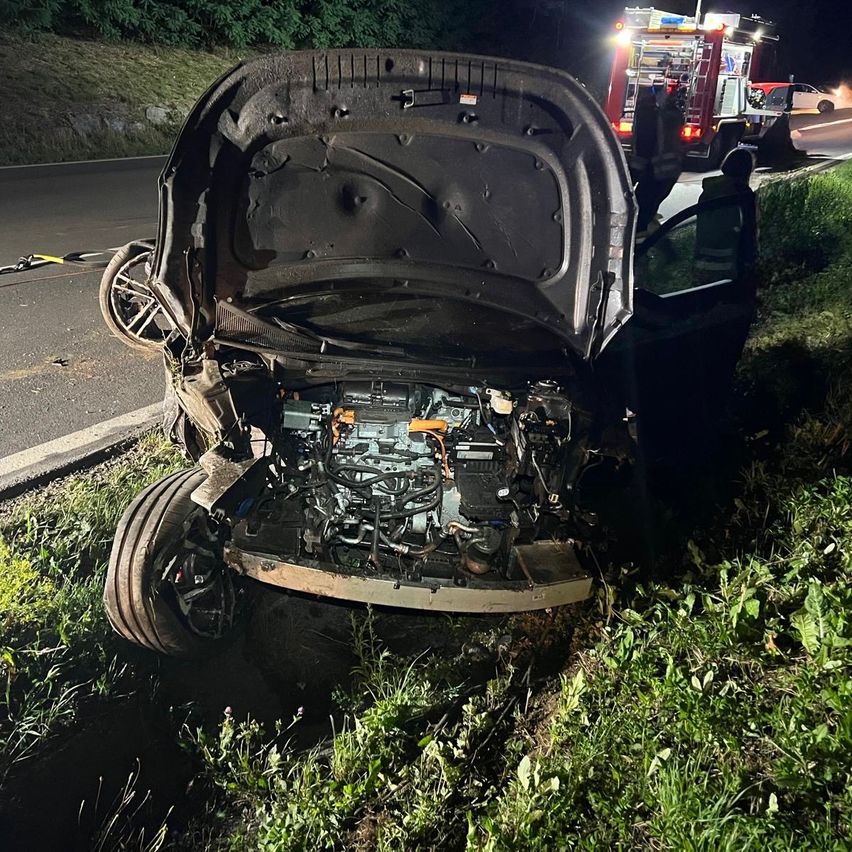 Ein Auto mit offenem Motorhaube liegt auf der Straßenseite, mit stark beschädigter Front. Rettungsdienste sind anwesend, und ein Feuerwehrwagen ist in der Nähe geparkt.