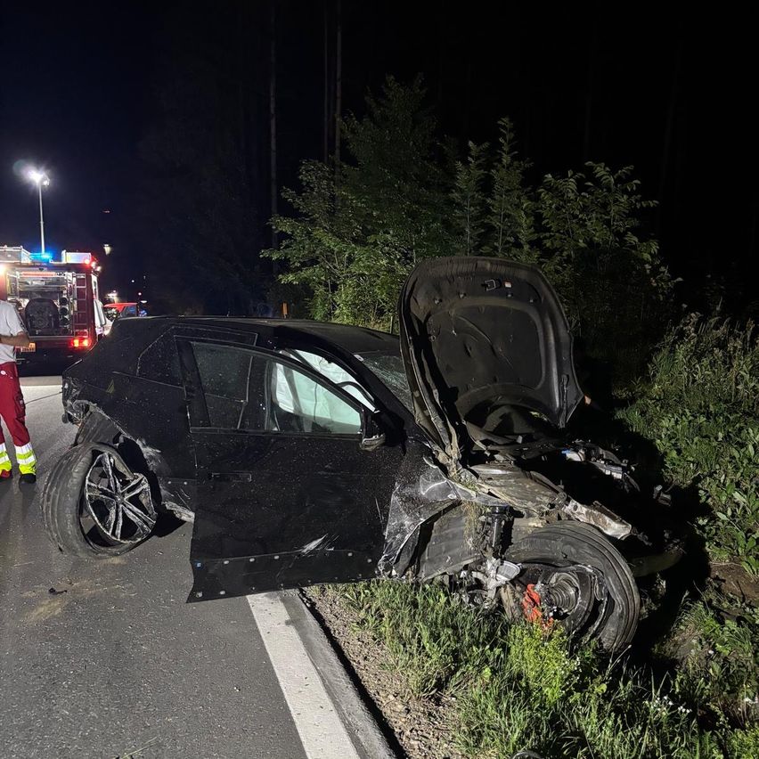 Ein schwarzes Auto ist nachts an der Straßenseite beschädigt. Die Motorhaube ist geöffnet und die Front ist stark beschädigt. Ein Feuerwehrwagen ist hinter dem Auto geparkt.