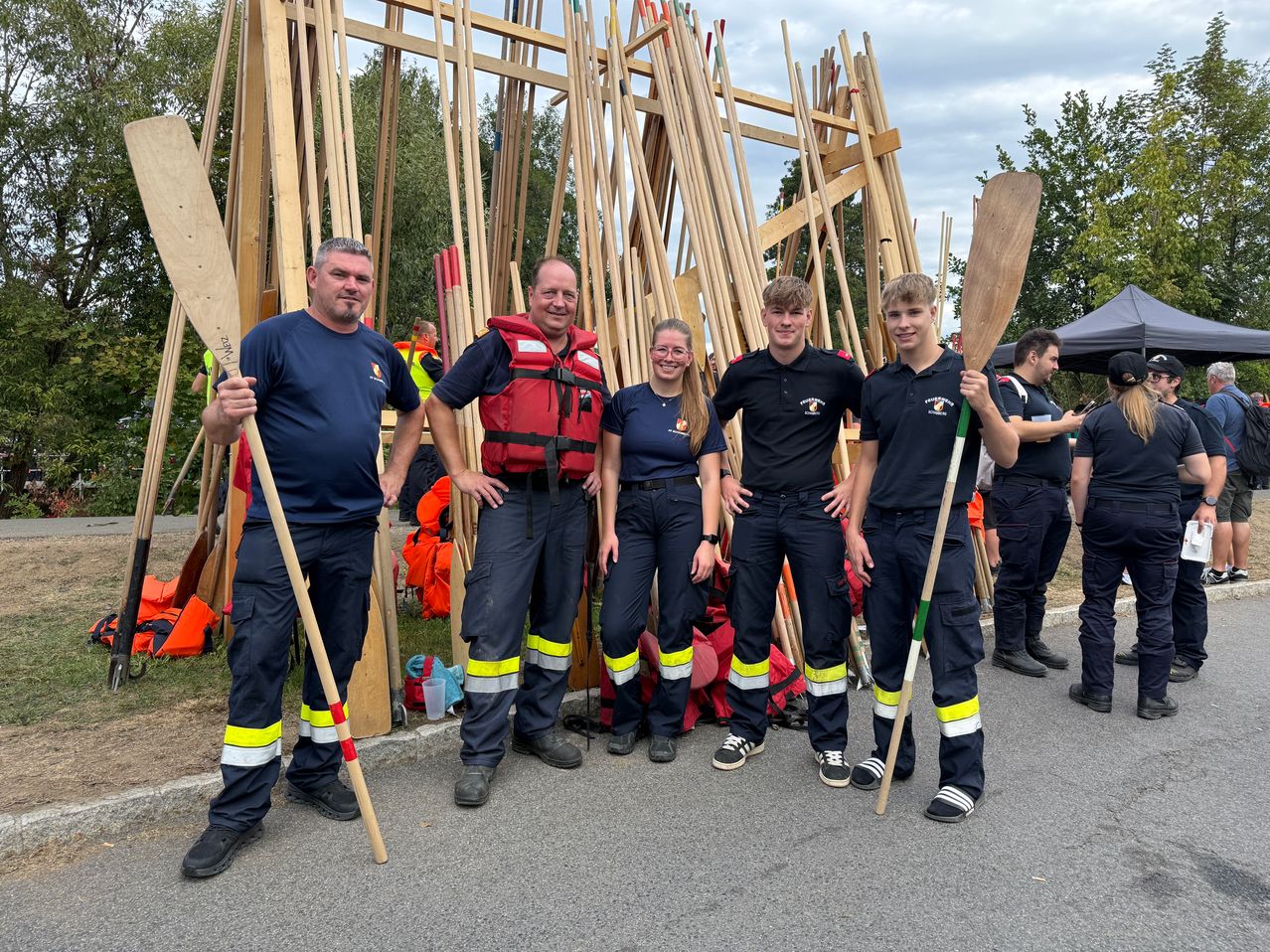 Five people in uniform stand in front of a large structure made of wooden poles, smiling for a photo.