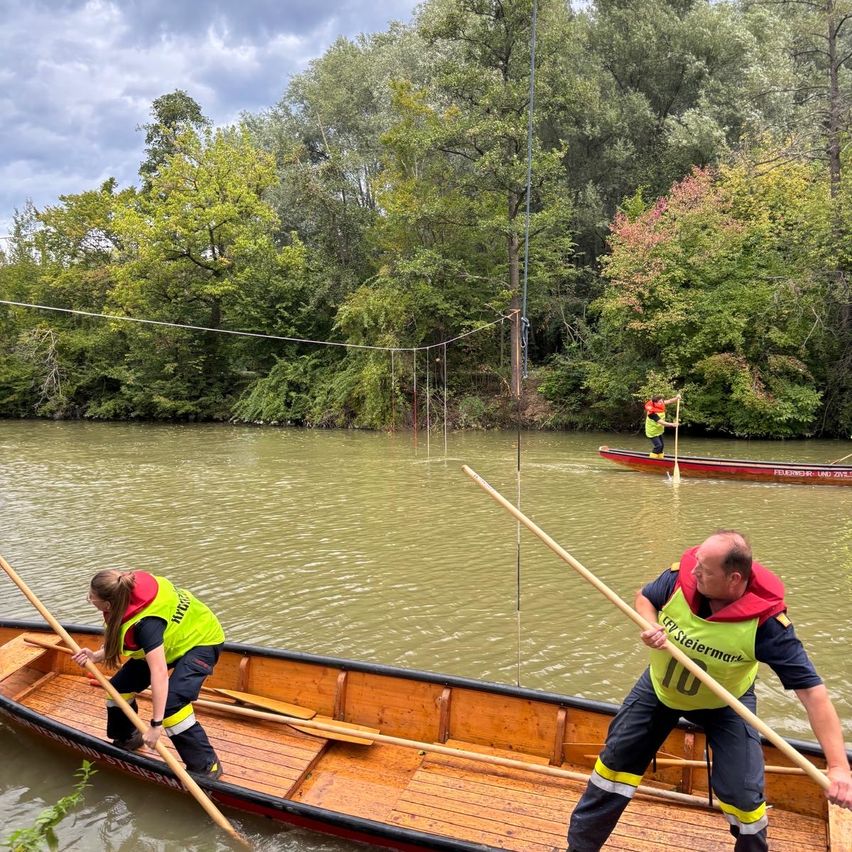 Two people in yellow vests paddle a wooden boat on a river with trees in the background. Another person stands in a separate boat.