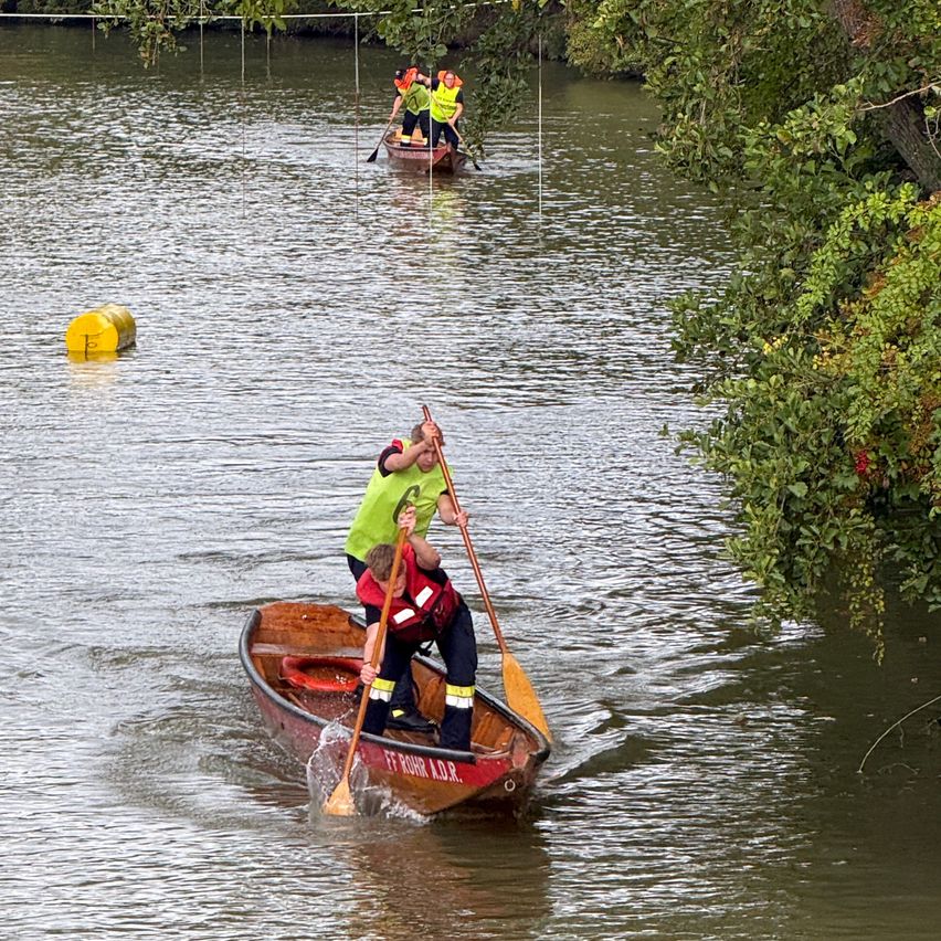 Two individuals in life jackets and green vests are rowing a red boat on a river. A yellow buoy floats nearby.