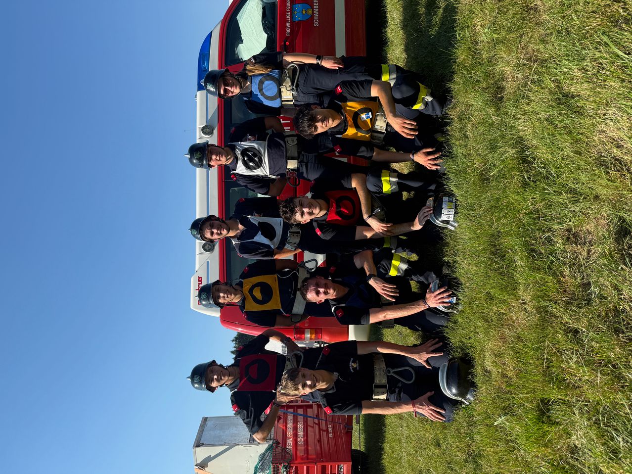 Eine Gruppe Feuerwehrleute posiert für ein Foto mit ihrem Feuerwehrwagen an einem sonnigen Tag.
