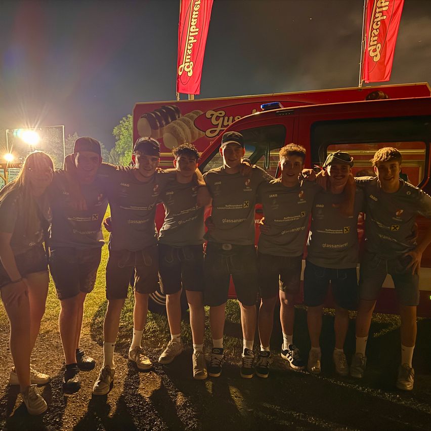 Eine Gruppe von Personen, möglicherweise ein Team, steht bei Nacht vor einem roten Fahrzeug. Sie tragen graue Shirts und Shorts. Hinter ihnen befinden sich rote Fahnen.