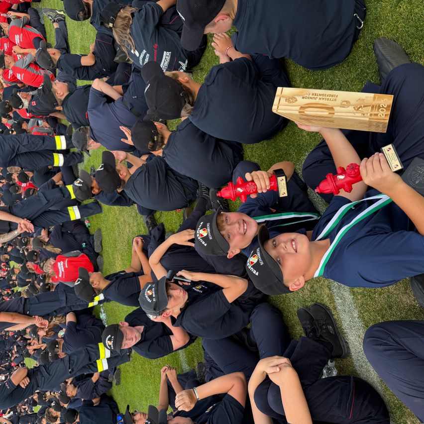 Eine Gruppe von Menschen in Uniform sitzt auf dem Rasen und hält Trophäen. Sie tragen Mützen und Turnschuhe, manche haben einen roten Feuerhydranten. Sie scheinen einen Sieg zu feiern.