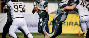 Drei American-Football-Spieler sind auf einem Feld. Der Spieler in der Mitte hält den Ball und bereitet den Wurf vor. Die anderen beiden Spieler versuchen, ihn zu blocken.