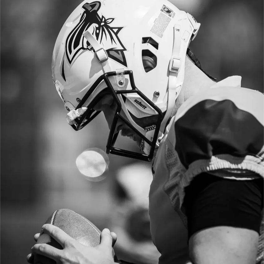 Ein Footballspieler in einem weißen Helm mit schwarzem und weißem Logo, der einen Football hält, schaut nachdenklich nach unten.