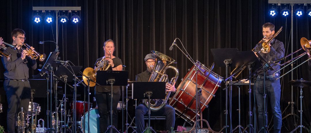Ein Musiker spielt die Trompete im Stehen, während ein anderer die Tuba im Sitzen spielt, auf der Bühne mit einem Schlagzeug. Beide spielen auf einer Bühne mit schwarzem Hintergrund.