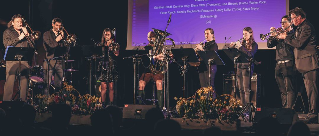 Ein Blechbläseroktett spielt auf der Bühne mit verschiedenen Instrumenten. Ein Bildschirm im Hintergrund listet die Namen der Musiker auf. Die Künstler tragen Freizeitkleidung und werden von Bühnenlichtern beleuchtet.
