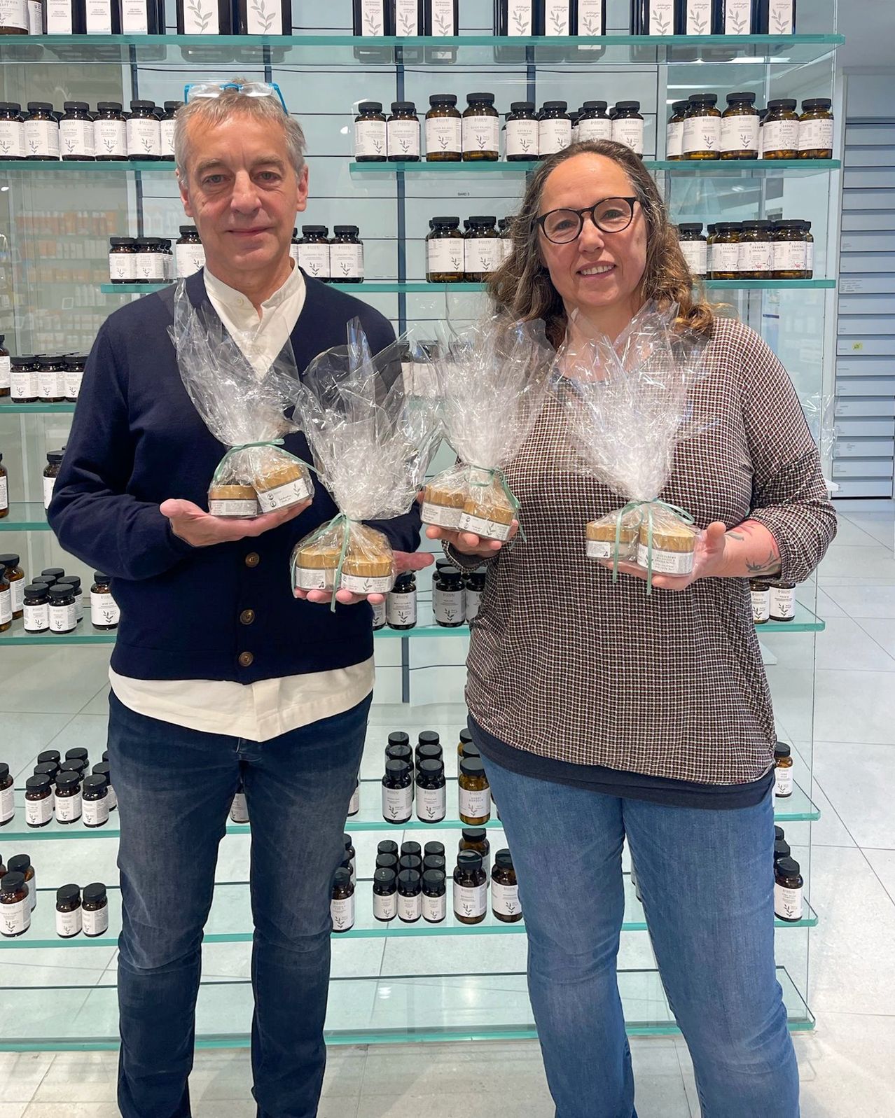 Zwei Personen stehen in einem Geschäft und halten lachend Pakete mit Keksen. Ein Regal mit verschiedenen Gläsern von Produkten steht hinter ihnen.