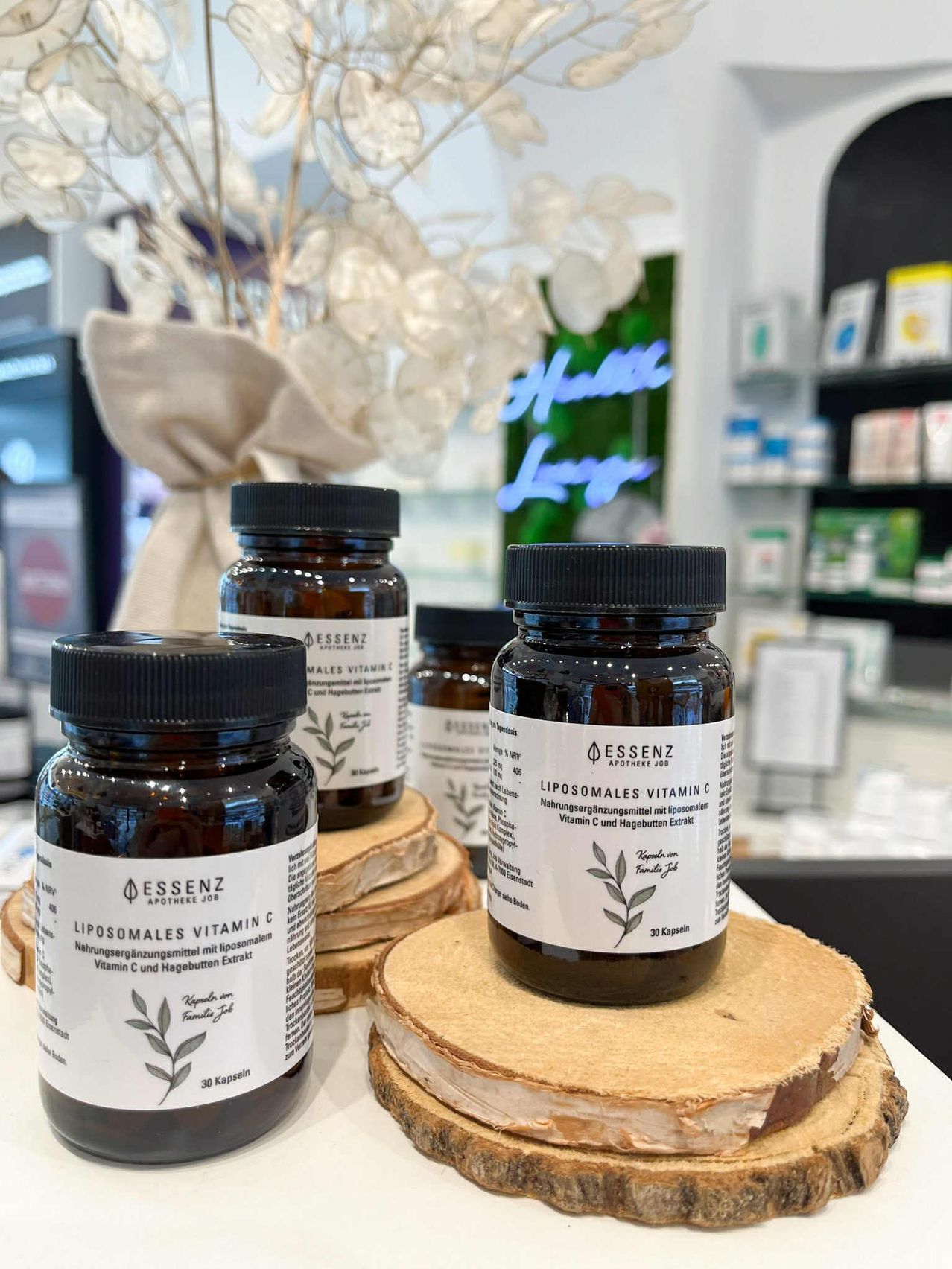 Mehrere braune Flaschen mit schwarzen Deckeln stehen auf Holzkreisen in einem Geschäft. Die Etiketten auf den Flaschen zeigen an, dass sie liposomales Vitamin C enthalten. Hinter den Flaschen steht eine weiße Pflanze in einem Beutel.