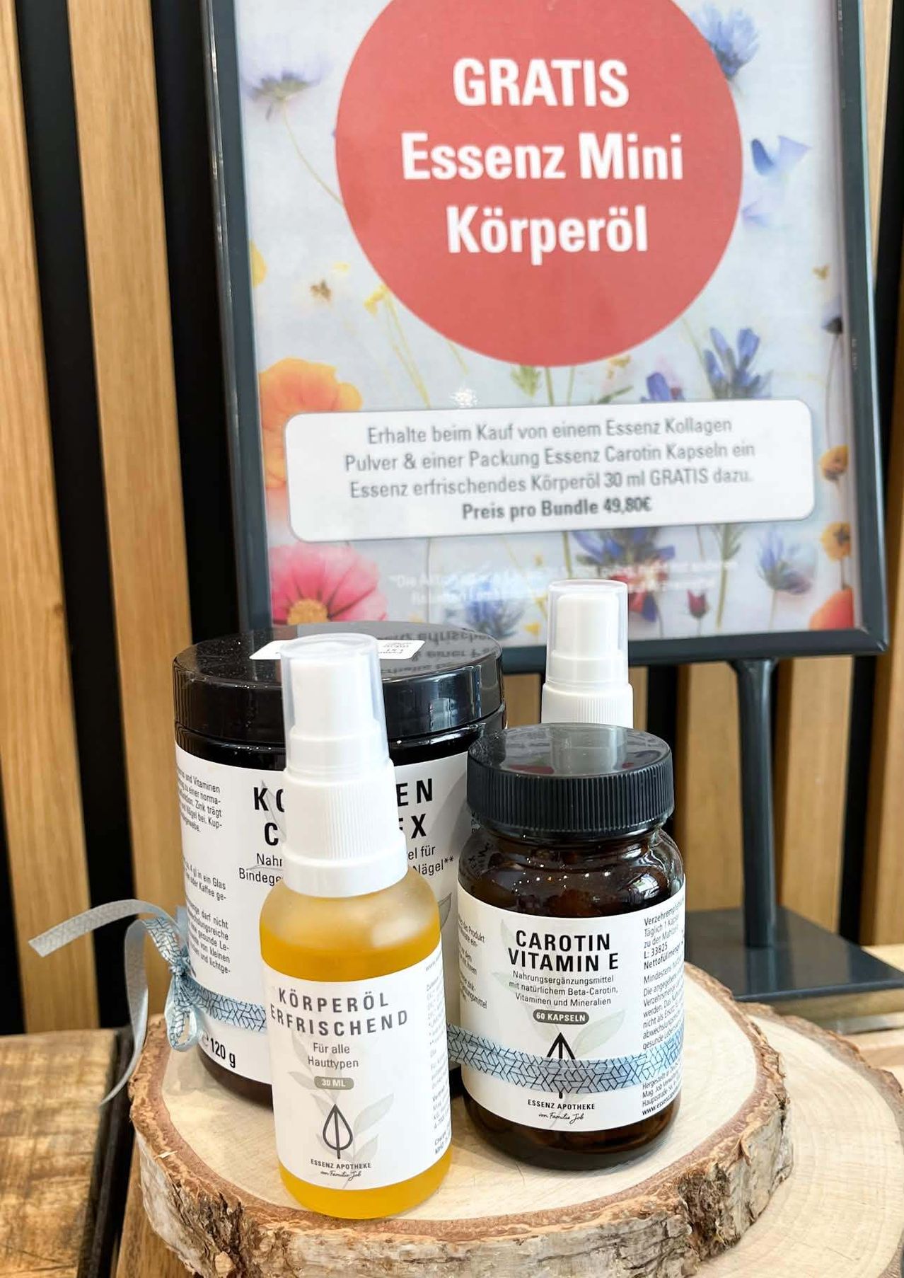 Ein hölzerner Tisch zeigt drei Flaschen und ein Glas mit Gesundheitsprodukten. Vorne steht eine Flasche erfrischendes Körperöl, ein Glas grünes Pulver und ein Glas Vitamine. Ein Plakat im Hintergrund trägt den Schriftzug 'Körperöl'.
