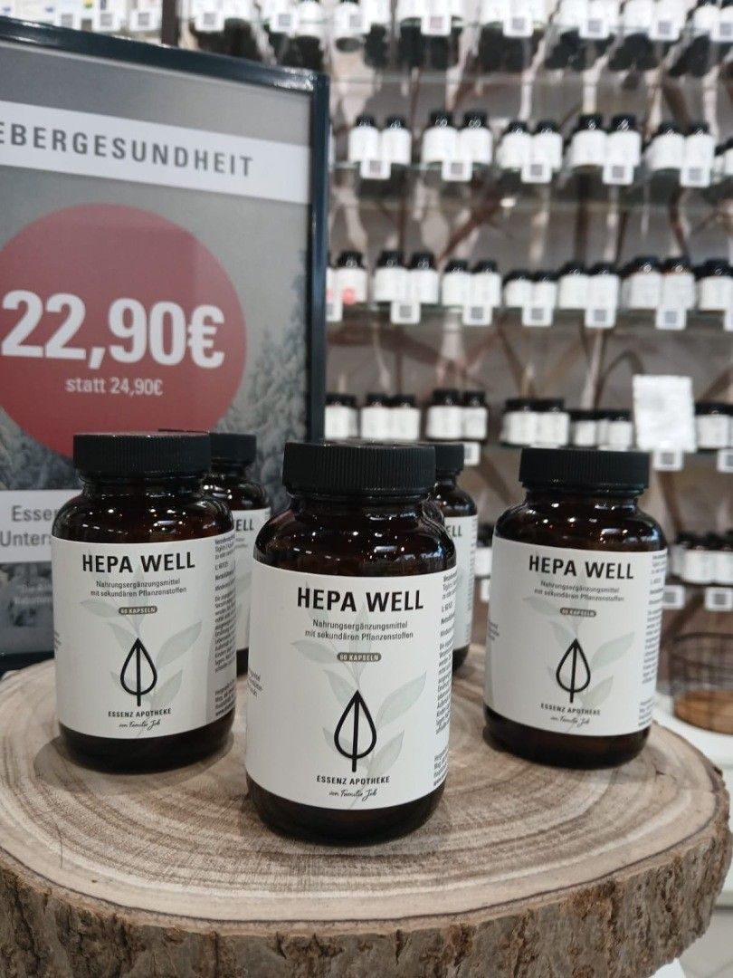 Drei Flaschen Hepa Well stehen auf einer Holzunterlage. Sie sind braun und weiß mit schwarzem Deckel. Dahinter befindet sich ein Regal mit mehreren weißen Flaschen.