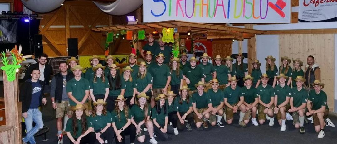 Eine Gruppe von Menschen in grünen Uniformen und Strohhüten posiert für ein Foto. Sie stehen vor einem Banner mit der Aufschrift Strohhatdisco. Einige Leute tragen Uhren und Turnschuhe.