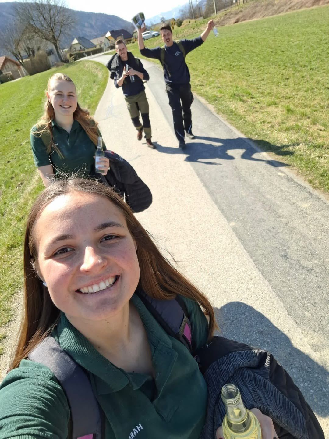 Drei Frauen in grünen Shirts gehen auf einem Betonweg. Die Frau vorne lächelt und hält eine Flasche. Dahinter gehen zwei weitere Frauen, eine hält eine Flasche und eine Jacke.