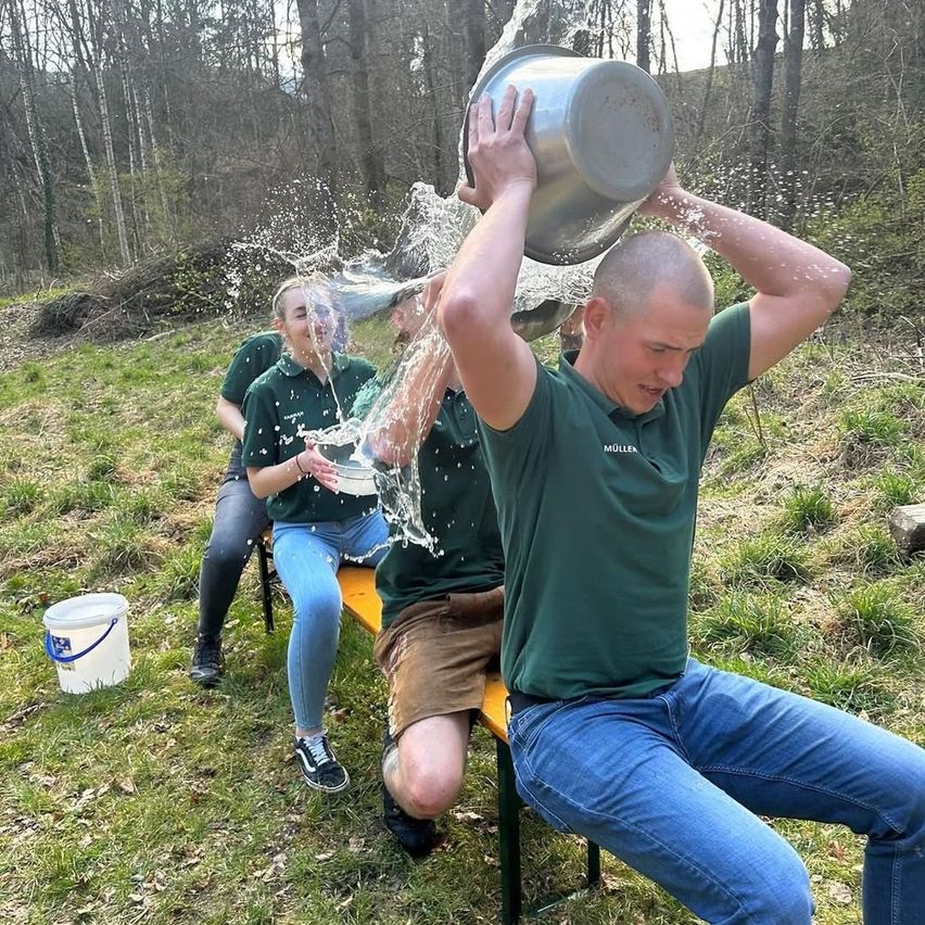 Ein Mann gießt sich Wasser aus einem Eimer über den Kopf, während er auf einer Bank sitzt. Hinter ihm beobachtet eine Frau, und ein weißer Eimer steht auf dem Boden.