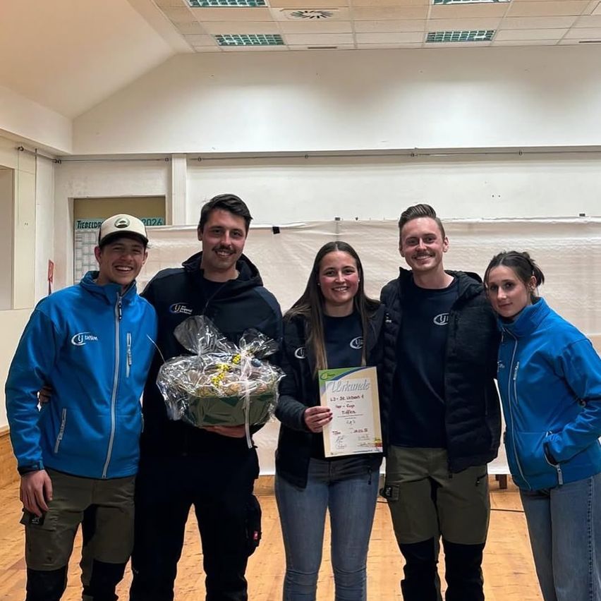 Fünf Personen, lächelnd, stehen in blauen Jacken in einem Raum. Eine Person hält ein Zertifikat und einen Korb mit Früchten. Der Raum hat Holzböden und weiße Wände.