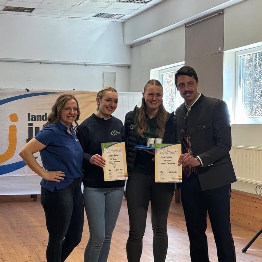 Vier Frauen und ein Mann posieren mit Zertifikaten und lächeln in einem Raum mit Holzböden und Bannern.