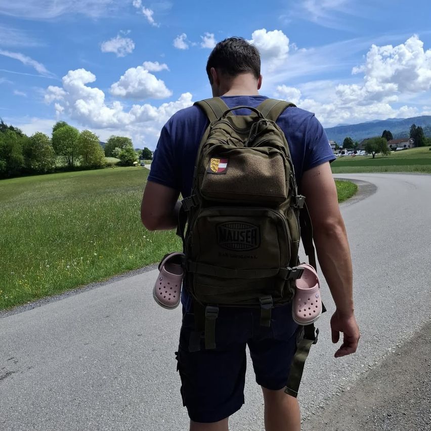 Ein Mann mit einem grünen Rucksack geht auf einer Straße. Seine Schuhe sind pink und hängen am Rucksack.