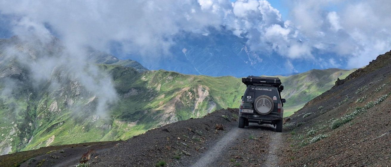 Ein Jeep fährt eine Bergstraße hinauf mit Wolken und Bergen im Hintergrund.