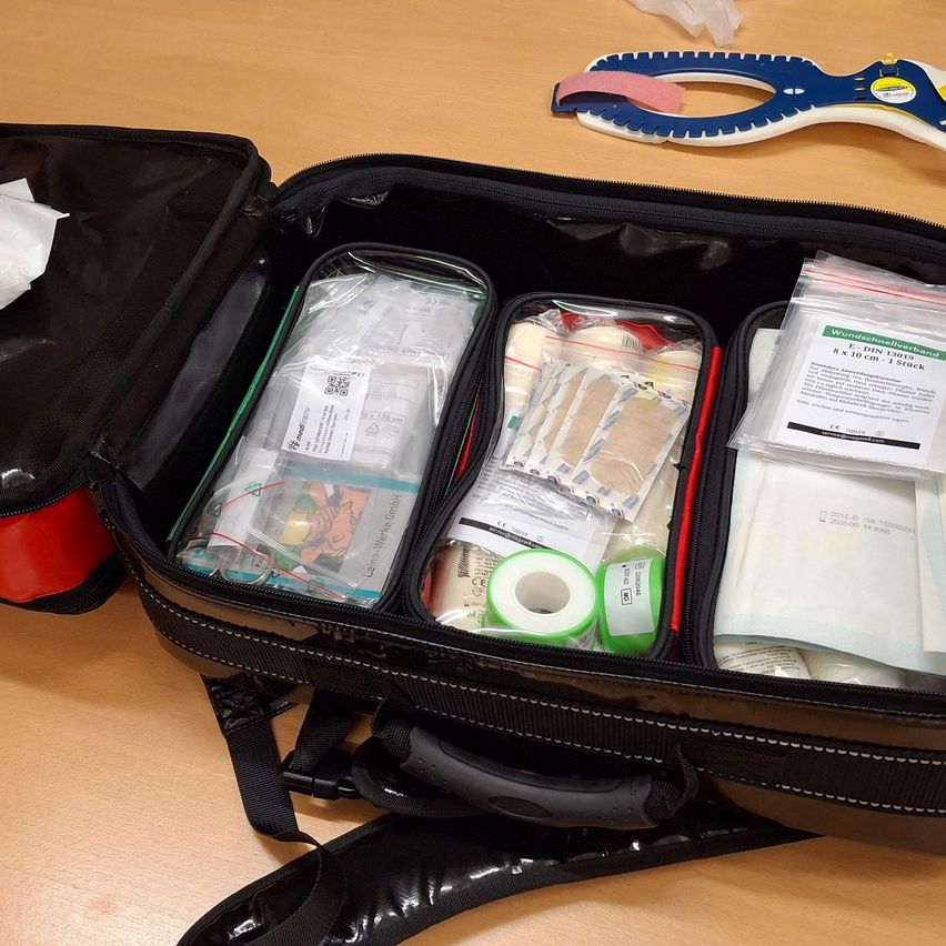 Ein offener schwarzer Koffer auf einem Holztisch enthält verschiedene medizinische Artikel, darunter Verbände, Klebebänder und eine Schiene.