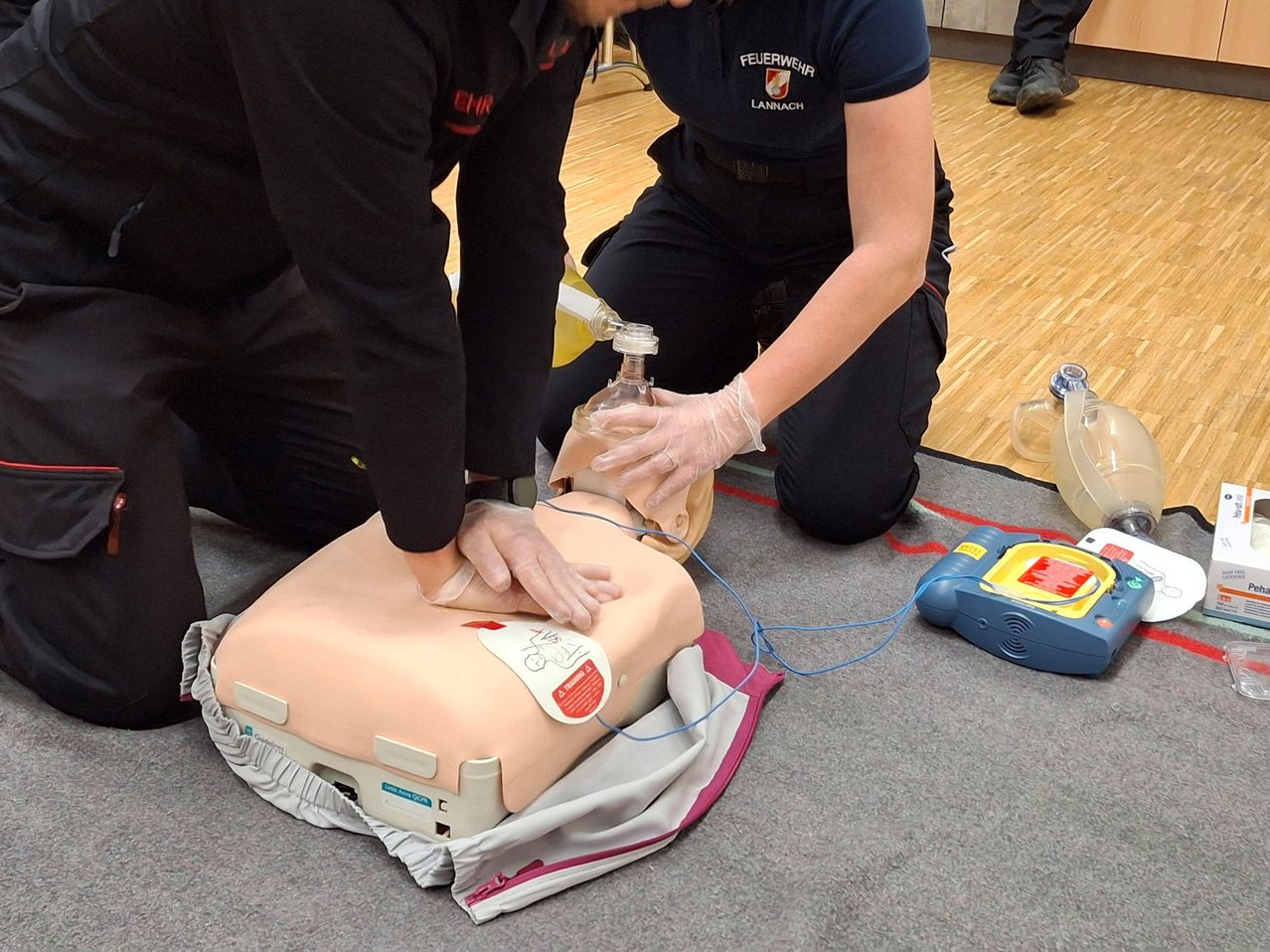 Zwei Feuerwehrleute üben CPR auf einer Puppe auf einem Teppich, mit einem Defibrillator in der Nähe. Ein Feuerwehrmann führt Brustkompressionen durch, während der andere den Defibrillator vorbereitet.