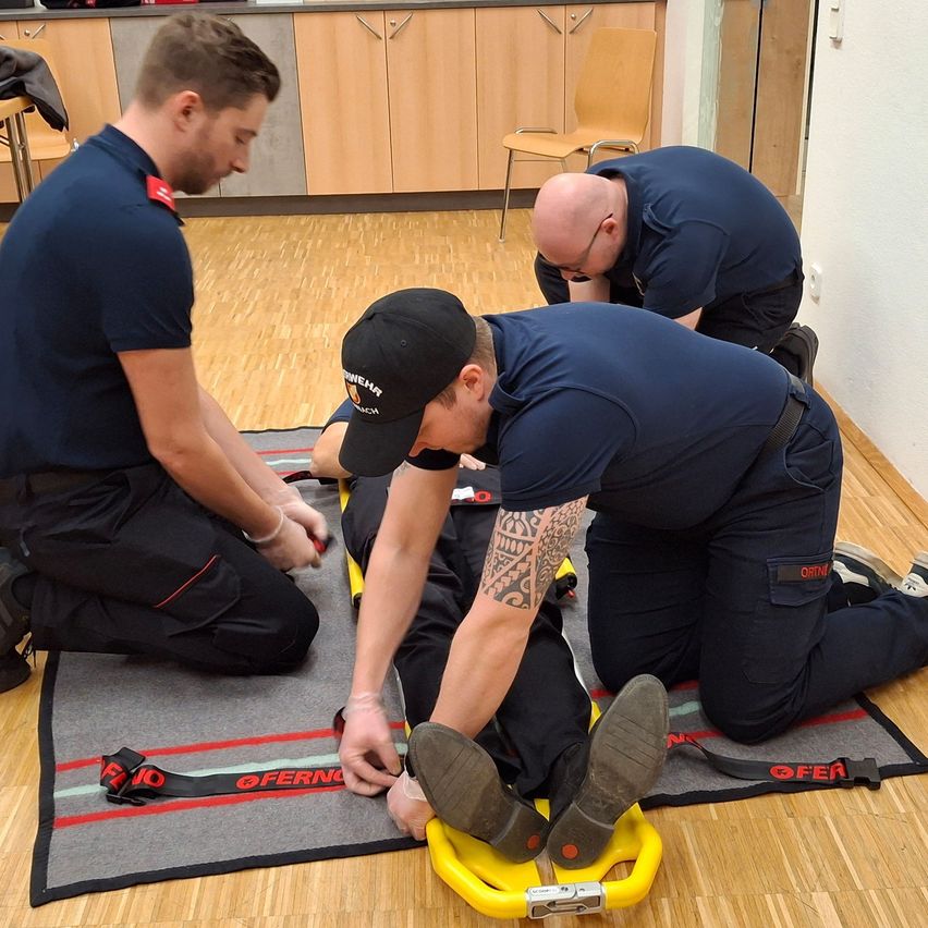 Drei Männer in blauen Uniformen führen eine Erste-Hilfe-Prozedur an einem Mann auf einer Matte durch. Der Mann liegt auf dem Rücken mit erhobenen Füßen. Ein Mann kniet auf der Matte und justiert eine gelbe Fußstütze.