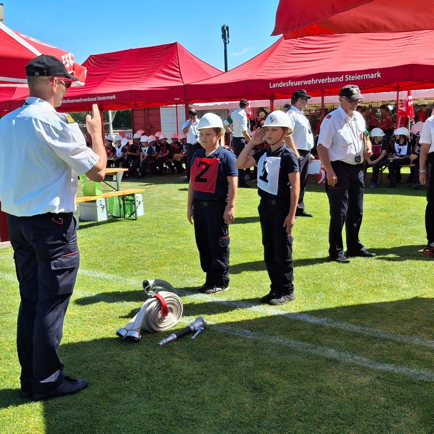 Zwei Kinder in Uniformen salutieren einem Mann in weißer Uniform auf einem Feld unter roten Zelten. In der Nähe liegen ein Feuerwehrschlauch und ein Werkzeug auf dem Boden.