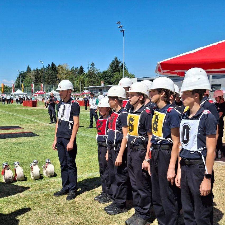 Eine Reihe von Kindern in weißen Helmen und nummerierten Uniformen steht auf einem Feld, mit einem roten Zelt im Hintergrund.