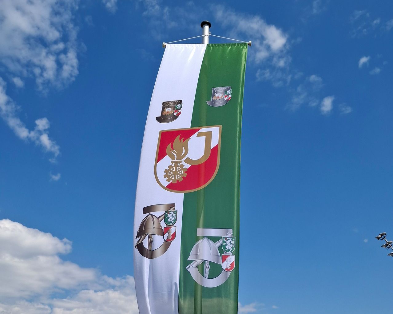 Ein Banner mit grünen und weißen Streifen, das verschiedene Embleme zeigt. Das obere Emblem zeigt ein Feuersymbol und ein Zahnrad, während das untere einen Helm und einen Schild zeigt. Es ist vor einem klaren blauen Himmel angebracht.