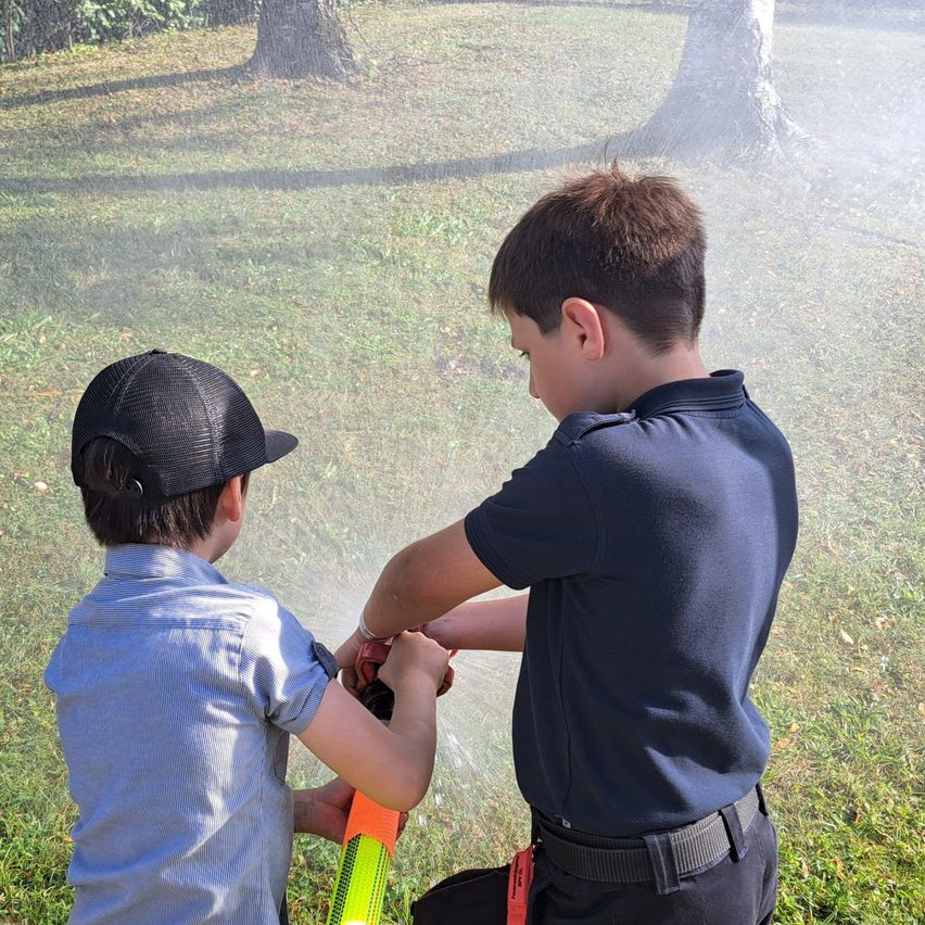 Zwei Jungen spielen auf einer Wiese mit einer Wasserpistole, einer trägt einen Hut und der andere ein blaues Hemd.