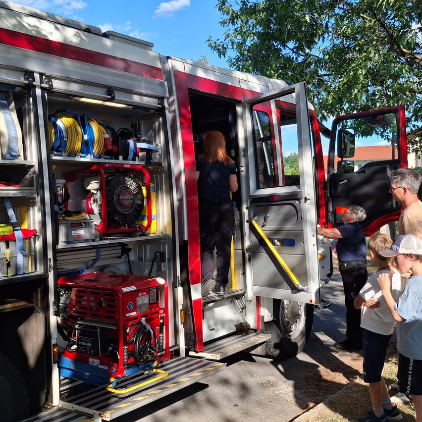 Eine Frau steigt in einen Feuerwehrwagen, während mehrere Kinder und Erwachsene beobachten, wobei Ausrüstung im Inneren des Wagens sichtbar ist.