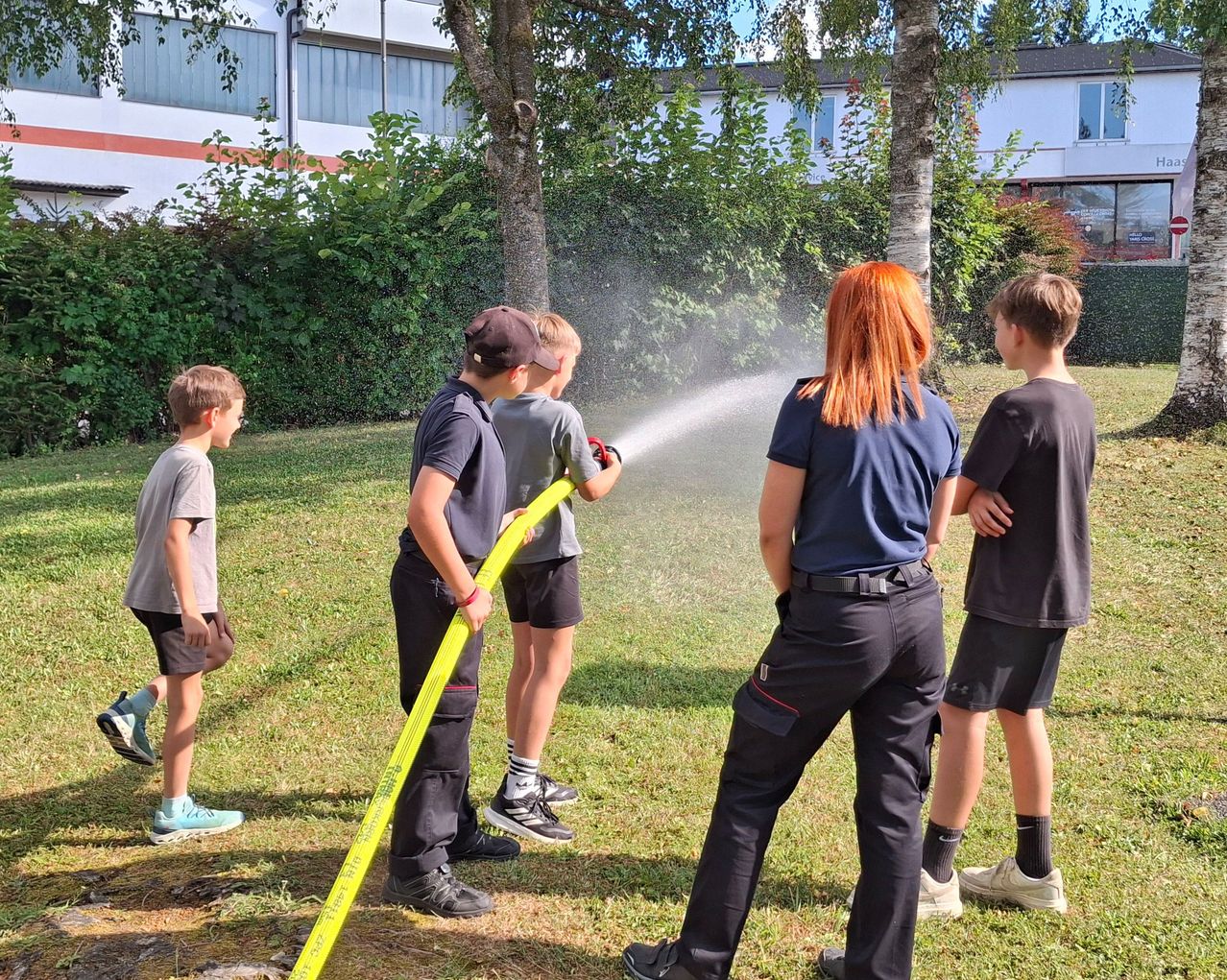 Eine Gruppe von Kindern und Erwachsenen ist draußen, wobei ein Erwachsener einen gelben Schlauch hält, der Wasser sprüht. Die Kinder beobachten das Wasser.