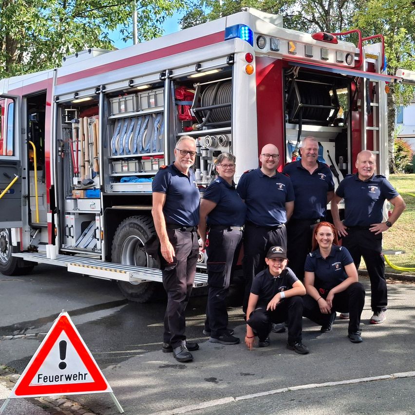 Eine Gruppe von Feuerwehrleuten steht vor einem Feuerwehrauto mit offenen Türen und zeigt verschiedene Ausrüstungsgegenstände im Inneren. Sie sind alle in Uniform gekleidet und lächeln für ein Foto.
