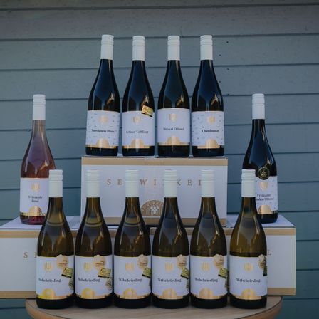 Eine Sammlung von Weinflaschen, die in zwei Reihen auf einem Holztisch angeordnet sind. Die oberste Reihe enthält verschiedene Weine, darunter Sauvignon Blanc, Grüner Veltliner und Chardonnay. Die untere Reihe besteht aus einer Reihe weißer Weinflaschen mit der Aufschrift 'Welschriesling'. Hinter den Flaschen befinden sich zwei Kisten, eine mit der Aufschrift 'SEWIKEI' und die andere mit einem ähnlichen Design.