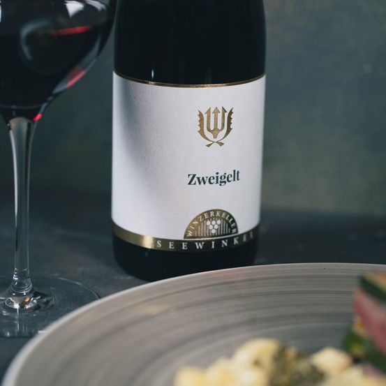 Ein Glas Rotwein steht neben einer Flasche Zweigelt-Wein. Die Flasche hat ein weißes Etikett mit goldenen Details. Ein Teller mit Essen steht in der Nähe.