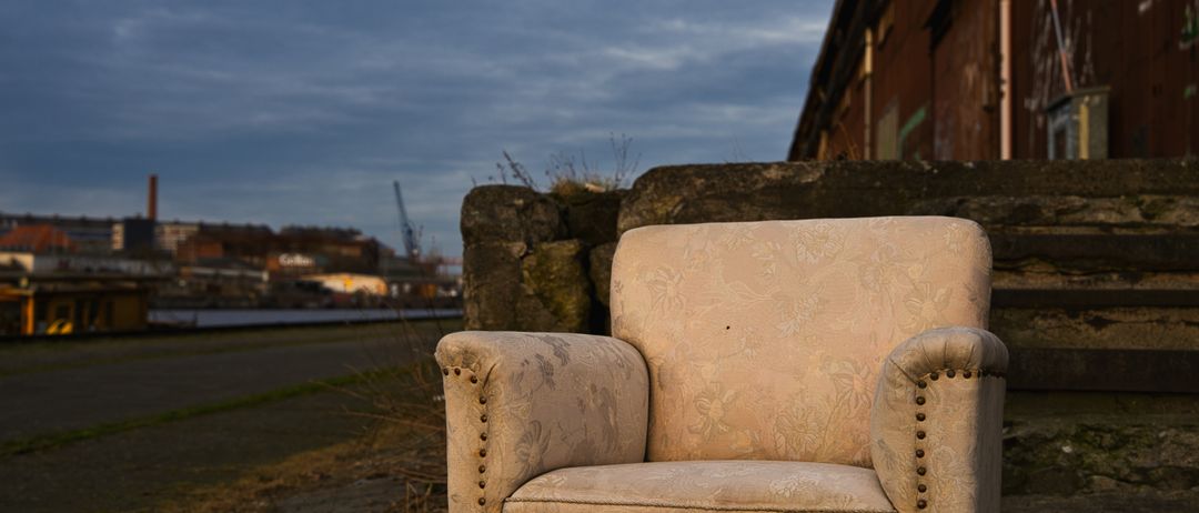 Ein alter Sessel mit Blumenmustern steht verlassen draußen, nahe einer Steinmauer und einem rostigen Gebäude. Der Himmel ist bewölkt.