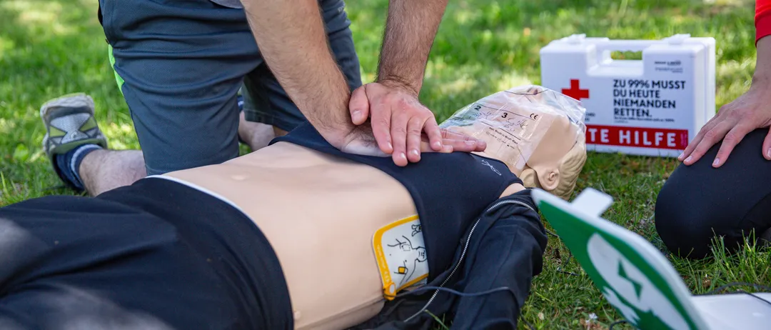 Eine Person übt CPR auf einem Mannequin, das auf Gras liegt. Ein Erste-Hilfe-Kasten ist in der Nähe.