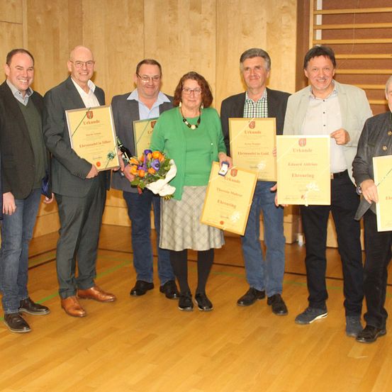Eine Gruppe von Menschen steht in einer Reihe, lächelt und posiert für ein Foto. Sie halten Urkunden und eine Person hat Blumen. Die Umgebung scheint ein Preisverleihungszeremonie zu sein.
