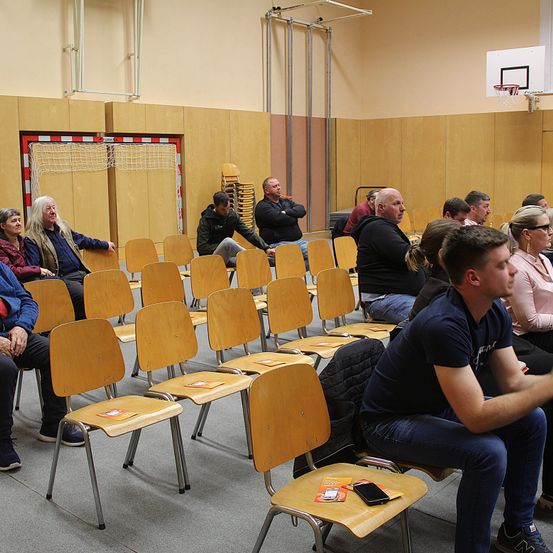 Eine Gruppe von Menschen sitzt in einem Saal mit in Reihen angeordneten Stühlen. Eine Basketballkorb ist an der Wand montiert. Ein Mann sitzt vorne mit verschränkten Armen.