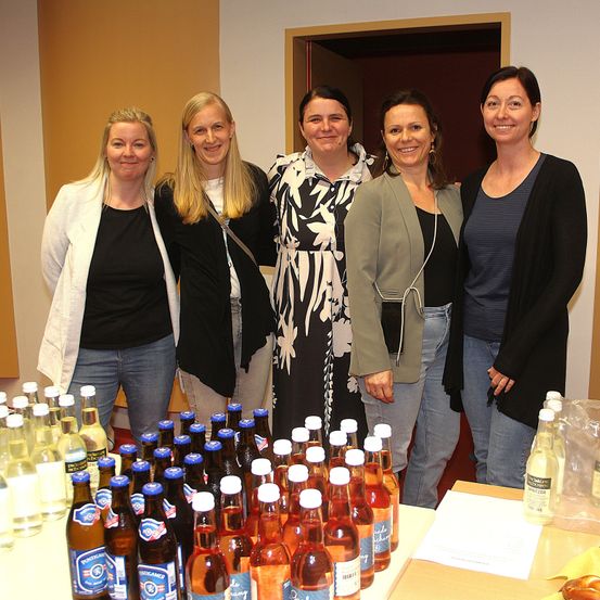 Fünf Frauen stehen vor einem Tisch mit verschiedenen Getränkeflaschen. Sie scheinen zu lächeln und für ein Foto zu posieren. Der Tisch enthält Flaschen mit Bier, Wein und Saft.