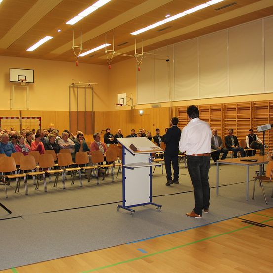 Ein Mann steht an einem Podium in einem Raum mit einer Menge Menschen, die auf Stühlen sitzen. Ein weiterer Mann steht in der Nähe, und ein Mikrofon befindet sich auf einem Tisch. Holzwände und Basketballkörbe sind im Hintergrund zu sehen.