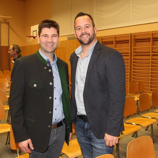 Zwei Männer stehen lächelnd in einem Klassenzimmer mit Holzstühlen. Der Mann auf der linken Seite trägt eine grüne Jacke und der Mann auf der rechten Seite trägt einen grauen Anzug. Hinter ihnen befinden sich Holzregale und ein Basketballkorb.