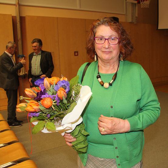 Eine Frau mit roten Haaren hält einen farbenfrohen Blumenstrauß in einem Raum, zwei Männer stehen hinter ihr, einer hält eine Kamera.
