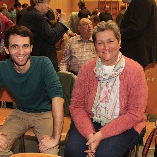 Eine Gruppe von Menschen sitzt und steht in einem Raum. Ein Mann in einem grünen Shirt lächelt und posiert für ein Foto. Neben ihm lächelt eine Frau in einem rosa Pullover und einer Schürze. Hinter ihnen sitzt ein Mann in einem karierten Hemd auf einem Stuhl.