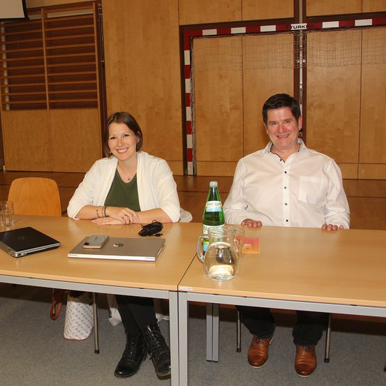 Zwei Personen sitzen an einem Tisch mit Laptops, einer Wasserkaraffe und einer Flasche. Eine Frau trägt einen weißen Schal und schwarze Stiefel, und ein Mann in weißem Hemd und braunen Schuhen.