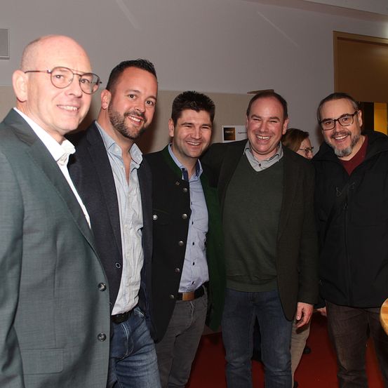 Fünf Männer stehen lächelnd für ein Foto zusammen. Sie befinden sich in einem Raum mit einem roten Teppich, weißen Wänden und einer Tür. Ein Mann hat eine Brille und einen Bart, ein anderer trägt eine Jacke und einen braunen Gürtel.