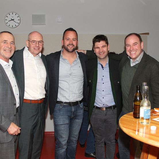 Fünf Männer in Anzügen posieren für ein Foto in einem Raum mit einer Uhr an der Wand. Flaschen stehen auf einem Holztisch vor ihnen.