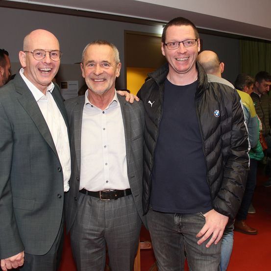 Drei Männer stehen lächelnd für ein Foto auf einem roten Teppich. Einer trägt eine Brille, ein anderer eine schwarze BMW-Jacke und der dritte einen Anzug. Im Hintergrund unterhalten sich weitere Personen.